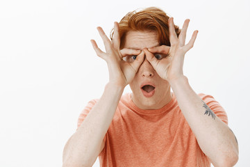 Portrait of charming carefree and childish redhead male with freckles, making goggles with fingers over eyes and gazing with opened mouth at camera, playing and fooling around joyfully