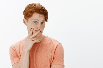 Studio shot of cute caucasian redhead with freckles and stylish hairstyle in orange t-shirt, touching lips and cheek and turning head, gazing with careless and indifferent expression at camera