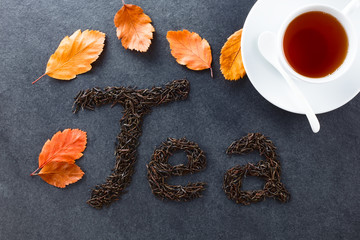 Loose black tea leaves spelling Tea on slate, autumn leaves and cup of black tea on the side, photographed overhead on slate (Selective Focus, Focus on the letters and the leaves)