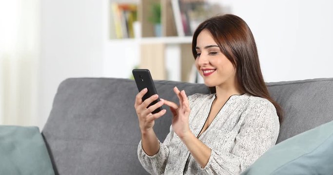 Happy woman browsing smart phone content sitting on a couch at home