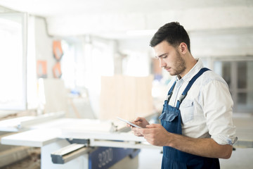 Young contemporary worker in overalls looking at touchpad display while browsing in workshop