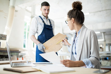 Fototapeta premium Two young contemporary engineers in workwear discussing characteristics of wooden board in workshop