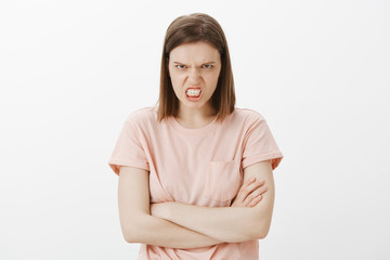 Fototapeta premium Hate, anger and negative emotions concept. Portrait of outraged and pissed woman, clenching fist grimacing from disdain, frowning eyebrows, looking from under forehead, holding hands crossed on chest
