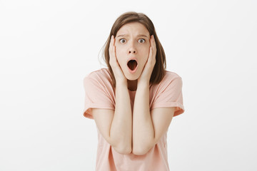 Shocked and stunned good-looking female model in pink t-shirt, dropping jaw, frowning and gasping holding hands on head, being displeased and troubled, standing disappointed over gray wall