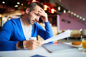 Fototapeta premium Pensive businessman with wireless earphones concentrating on paperwork while working in cafe