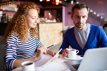 Two young casual colleagues sitting in cafe in front of laptop and working over project
