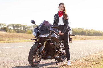 Fototapeta na wymiar Photo of active female motocyclist sits on motorbike, wears fashionable leather jacket, red bandana on neck, poses in unknown place outdoor, feels free and relxed. People and travelling concept