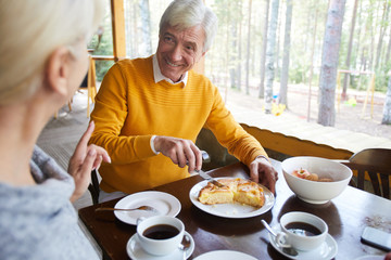 Happy senior man cutting apple pie while talking to his wife by tea in cafe