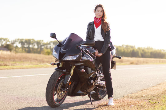 Photo of active female motocyclist sits on motorbike, wears fashionable leather jacket, red bandana on neck, poses in unknown place outdoor, feels free and relxed. People and travelling concept