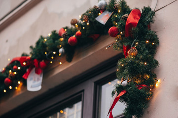 Stylish christmas decorations, garland lights and fir branches with red ornaments on window in european city street. Festive decor and illumination in winter holidays city center