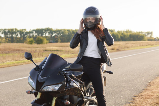 Horizontal view of stylish confident female biker dressed in fashionable clothes, sits on motorbike, wears helmet, covers long destination, poses outdoor. Motorcyclist enjoys outdoor travel.