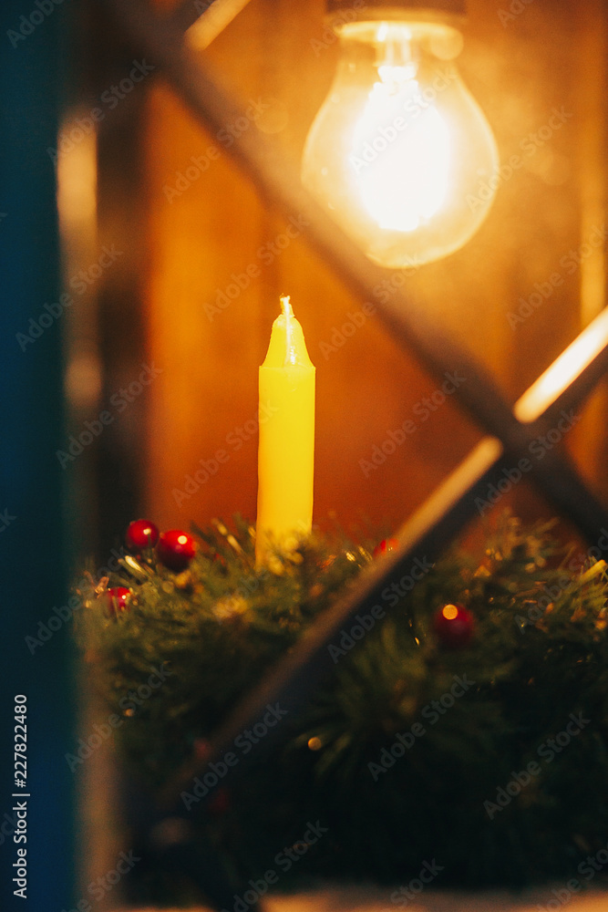 Wall mural Christmas candle in christmas wreath with berries on  window  in european city street. Wax candle under bulb lights. Simple Festive decorations and illumination in winter holidays