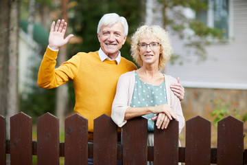 Hospitable mature couple standing by fence while friendly man waving hand to their neighbours