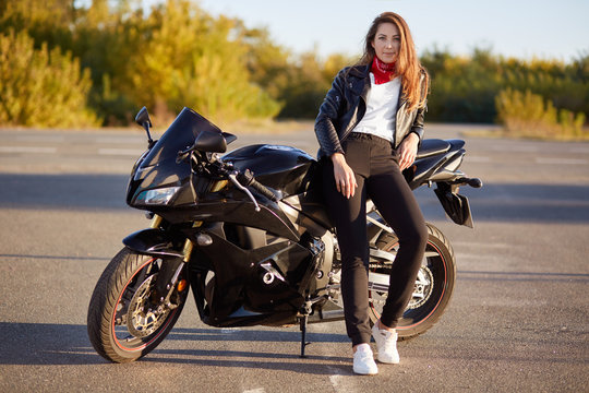 Photo of attractive woman has real adventure outdoor, stands near motorbike, dressed in bikers outfit, enjoys sunny day and calm atmosphere. People and motorcycling concept. Its time for ride
