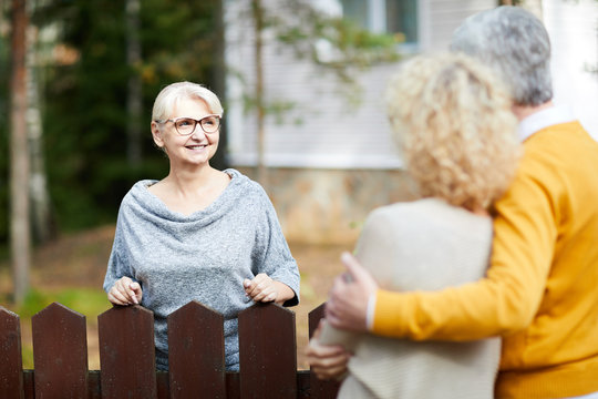 Blonde mature woman in eyeglasses and grey pullover looking at her neighbours during talk through fence
