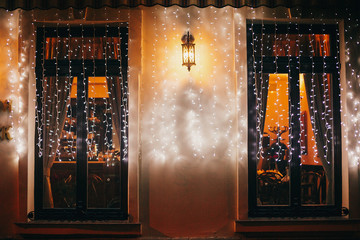 Stylish christmas decorations, garland lights and lantern on window in european city street....