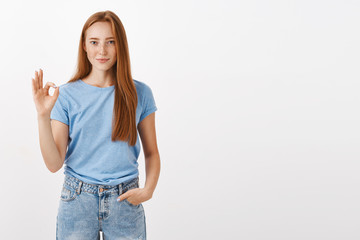 Obraz premium Ok it is deal. Portrait of good-looking satisfied and happy redhead female student in blue t-shirt holding hand in pocket showing okay or perfect gesture with fingers smiling liking or accepting plan