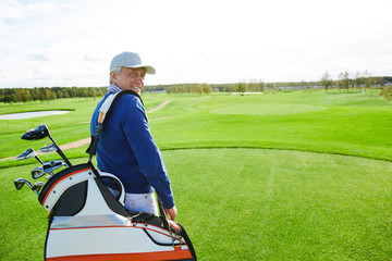 Happy senior man with bunch of golf clubs in bag looking at camera while walking down green field