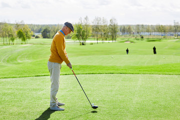 Fototapeta premium Senior man in casualwear standing in the middle of green field and going to hit golf ball