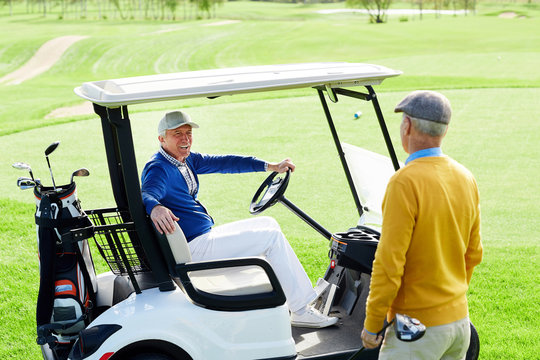 Aged man sitting in golf car and talking to his buddy standing on green field