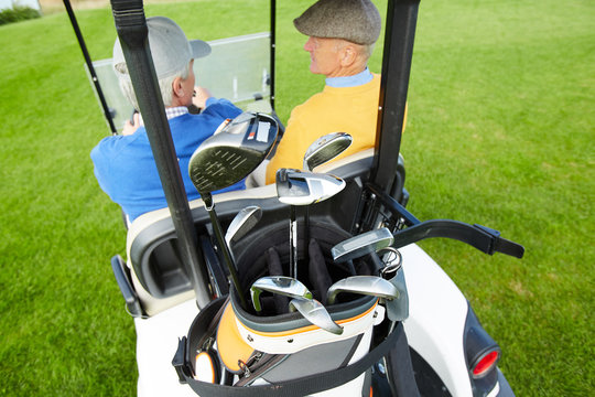 Back view of golf car with bunch of golf clubs and two mature men talking in cab