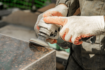 a man in work clothes and gloves polishes a marble stone with an angle grinder. grinding stone. manufacturing of monuments. marble slab