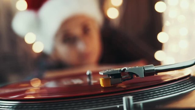 Christmas. Vinyl record player. Blurred women in a Christmas hat with red hair, listening to a vinyl record and singing a Christmas melody in the background with bright bokeh of Christmas lights