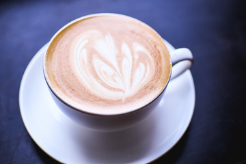 White cup of cappuccino with an abstract drawing on a brown coffee foam. Elegant coffee art. Concept background of cafe, rest, leisure, and romantic settings. Deep black background