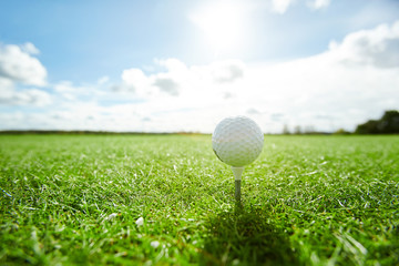 White golf ball on tee on vast green play field with cloudy sky and sunshine above