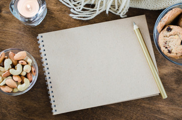 Flat lay of blank brown notebook with knitted blanket and snacks on wooden table.