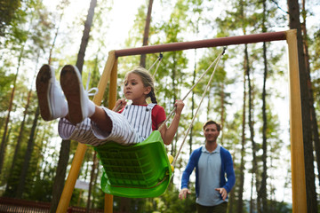 Little girl sitting on swing while her father pushing her from behind during chill in park