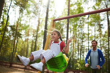 Cute little girl enjoying chill with her dad while sitting on swing in natural environment