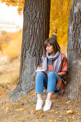 young woman in fall forest composes poems or the text songs. Romantic mood concept. Writes in a notebook. Brunette woman in autumn park with fashionable plaid coat and scarf.