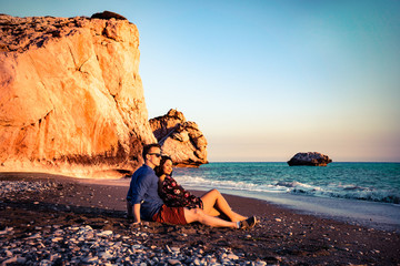 Young loving couple sitting together at the beach in front of the aphrodite rock in Cyprus Greece...