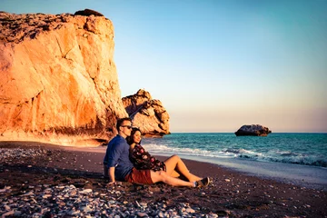 Poster Young loving couple sitting together at the beach in front of the aphrodite rock in Cyprus Greece during sunset. © Saga_bear