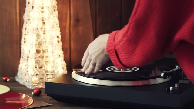 A young woman in a red knitted winter pullover puts a black vinyl record on a turntable, a stylus a needle and listens to music against the background of a glowing Christmas tree and a cozy New Year's