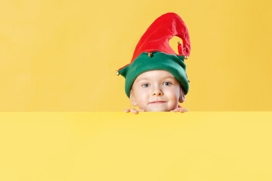Little girl in a red Christmas elf hat on a yellow background. The child is peeping from behind the sheet with a copy space.