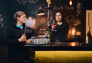 Handsome guy in hoodie smokes a hookah and seductive brunette woman drinks wine at nightclub or bar.