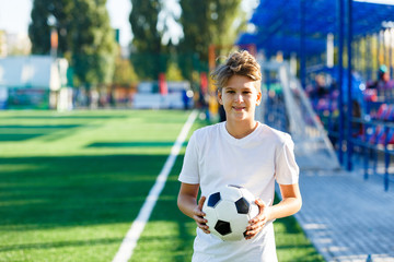 cute young boy in white blue sportswear holds classical black and white football ball on the green grass of the stadium field. Soccer game, training, hobby concept. with copy space 