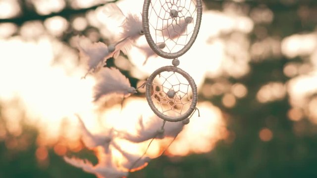 Slow motion amulet dreams catcher waving in a light wind against the backdrop of a bright sunset and a bokeh of city lights. White light feathers rise in the wind. The disc of the setting sun. Ethnic