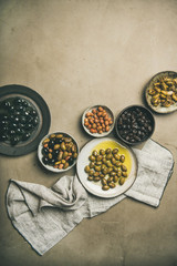 Flat-lay of various kinds of Mediterranean pickled olives in plates over grey concrete table background, top view, copy space, vertical composition. Mediterranean meze appetizer