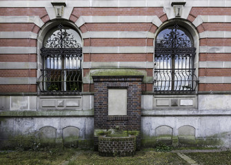 old rusty water fountain bluild from red bricks near a pink gray brick wall, with two windows with decorated bans, Lubeck, Germany