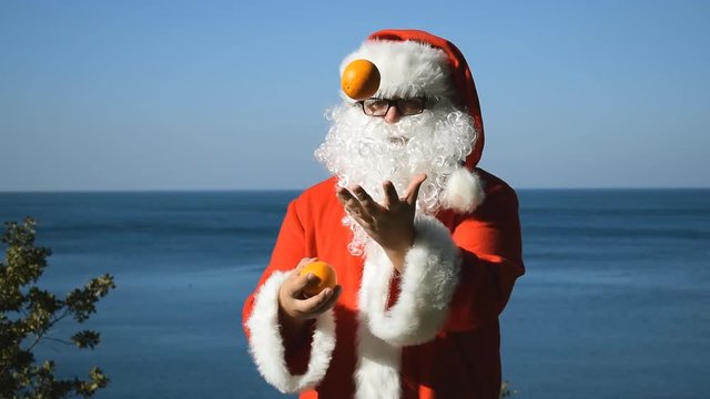 A man in a Santa Claus costume juggles tangerines on the seashore. Travel and vacation