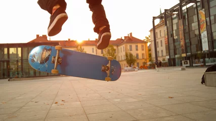 Rolgordijnen LOW ANGLE: Blue skateboard flipping underneath the young skateboarder's feet. © helivideo