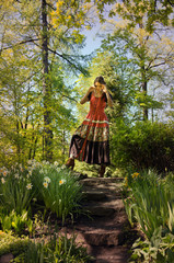 woman in a red boho style dress walks among blooming flower beds in a forest garden