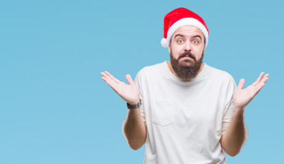 Young caucasian hipster man wearing christmas hat over isolated background clueless and confused expression with arms and hands raised. Doubt concept.