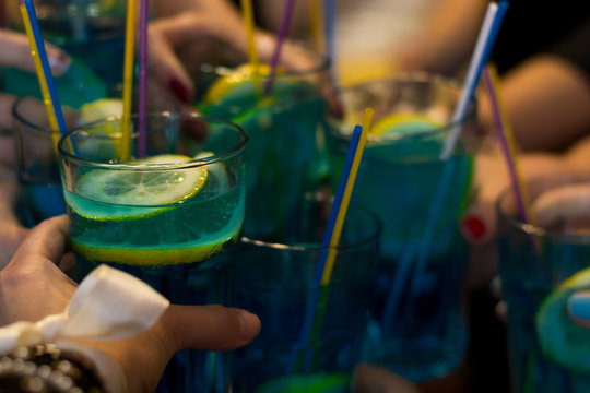 Clinking glasses full of blue cocktails with lemon slices and colorful tubes. Process of celebrating hen party. Girl becomes wife. Girls friends party in cocktail bar. Alcohol beverages in hands.