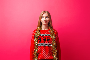 an emotional girl in a Christmas sweater and with tinsel on her neck, with a cunning expression, thought about something, isolated on a red background.