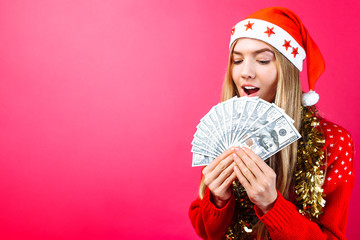 Emotional girl in a red sweater and Santa Claus hat, in admiration holding money on a red...