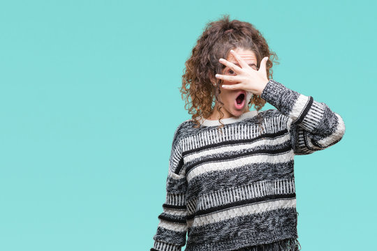Beautiful brunette curly hair young girl wearing glasses over isolated background peeking in shock covering face and eyes with hand, looking through fingers with embarrassed expression.